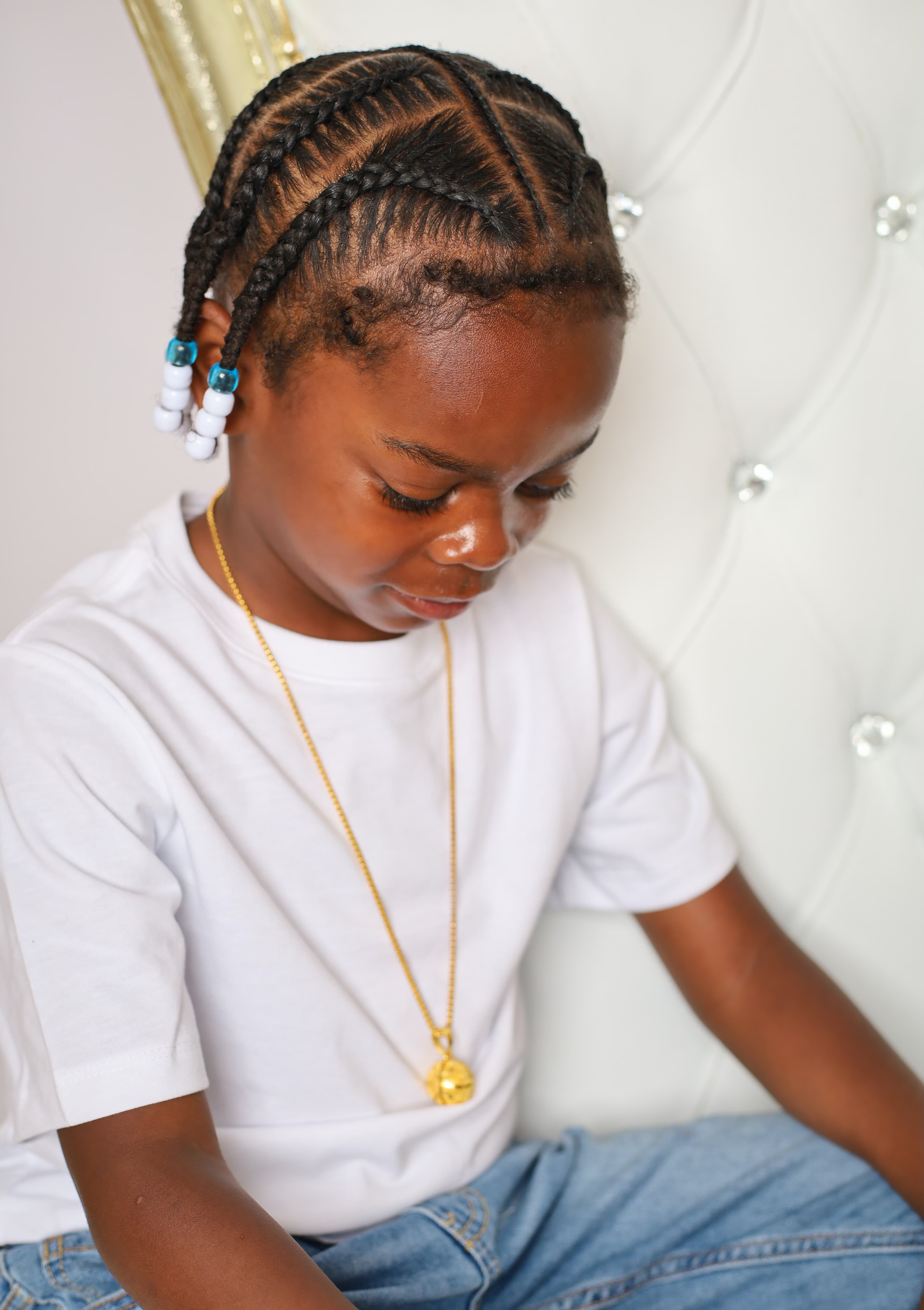 Young Black boy with beaded braids and a gold basketball necklace looking down thoughtfully.