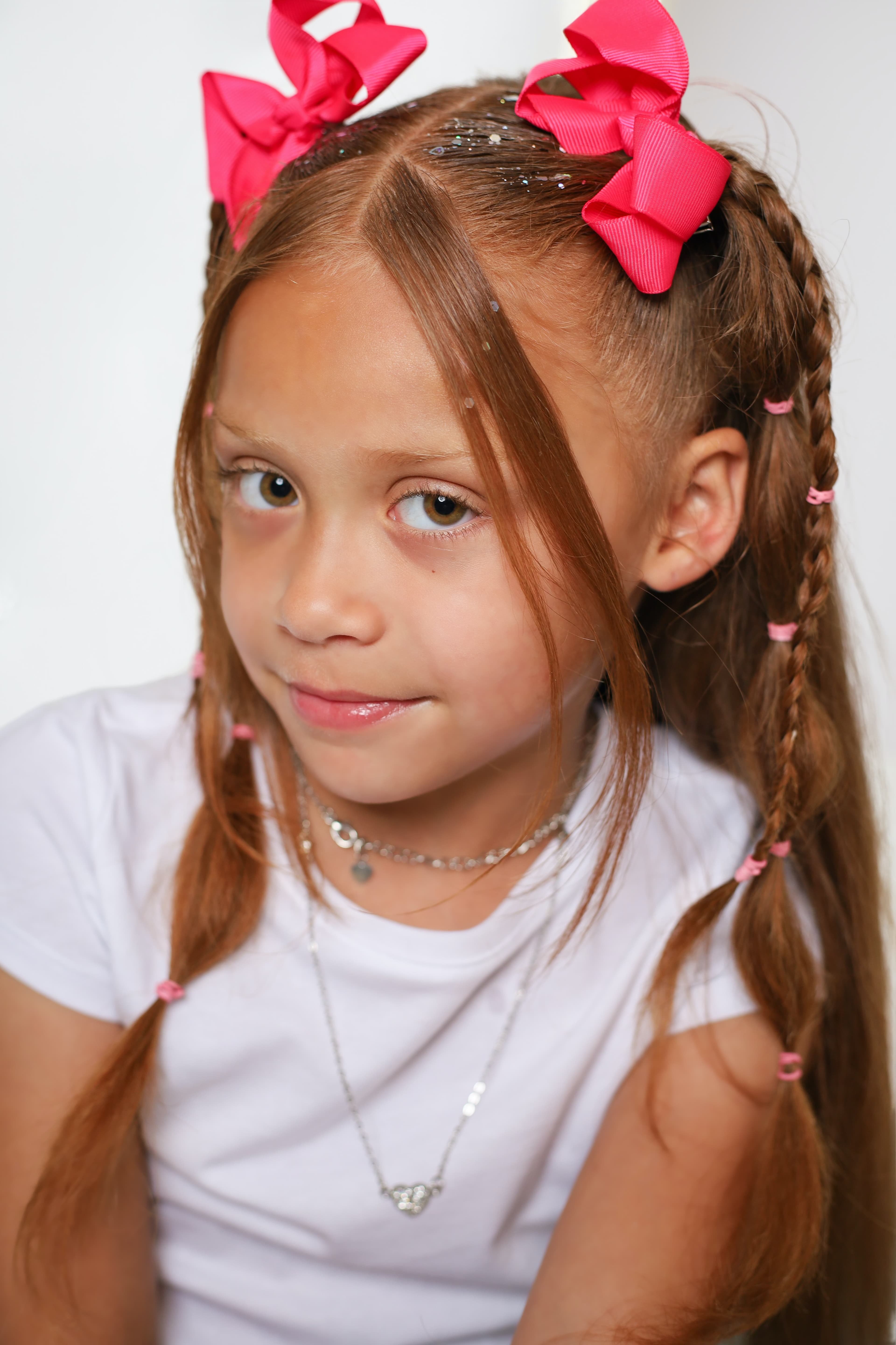 Young girl with reddish hair, pink bows, and glitter, wearing a white shirt and necklaces.