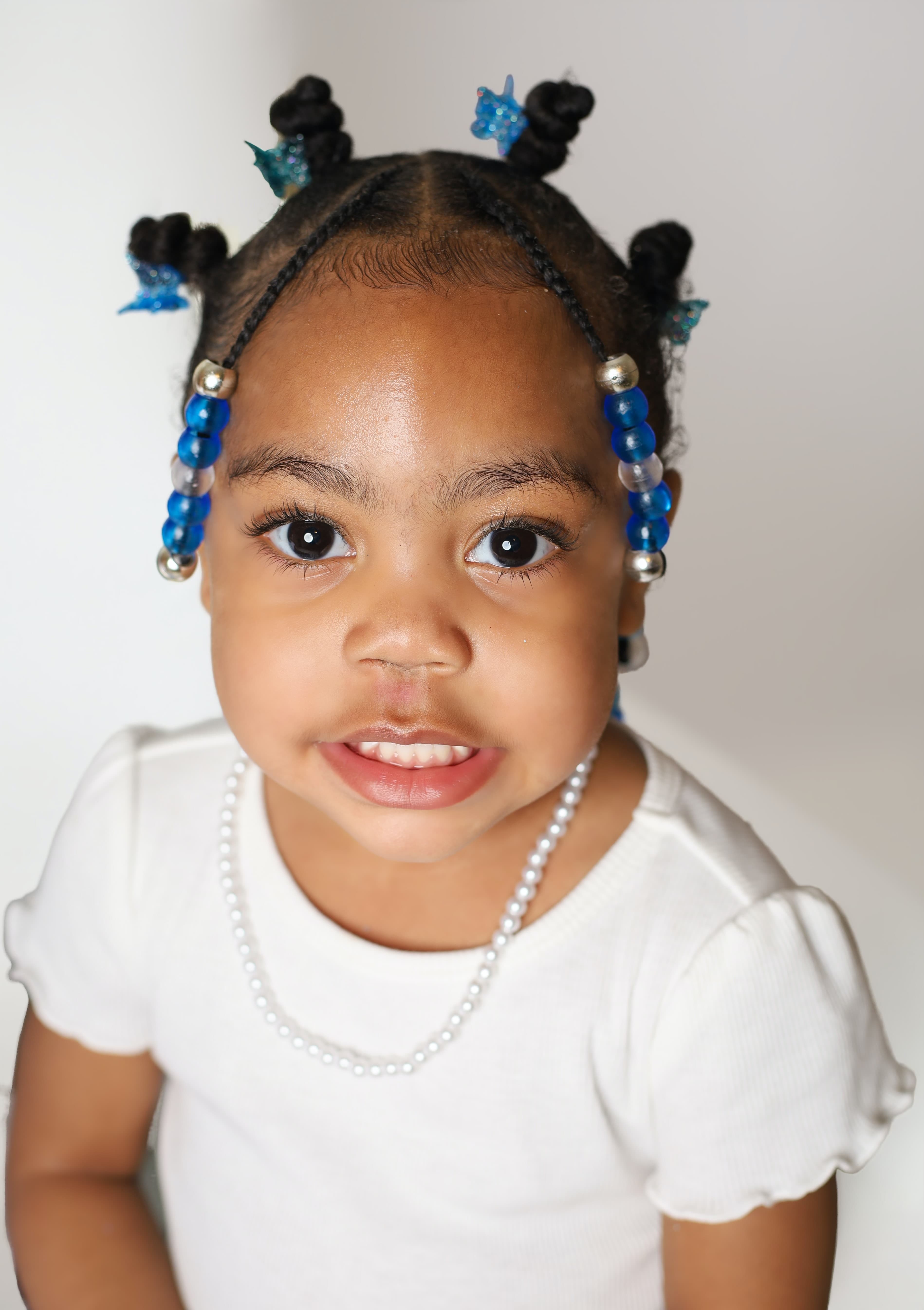 Smiling young Black girl with blue hair beads, butterfly clips, and a pearl necklace.